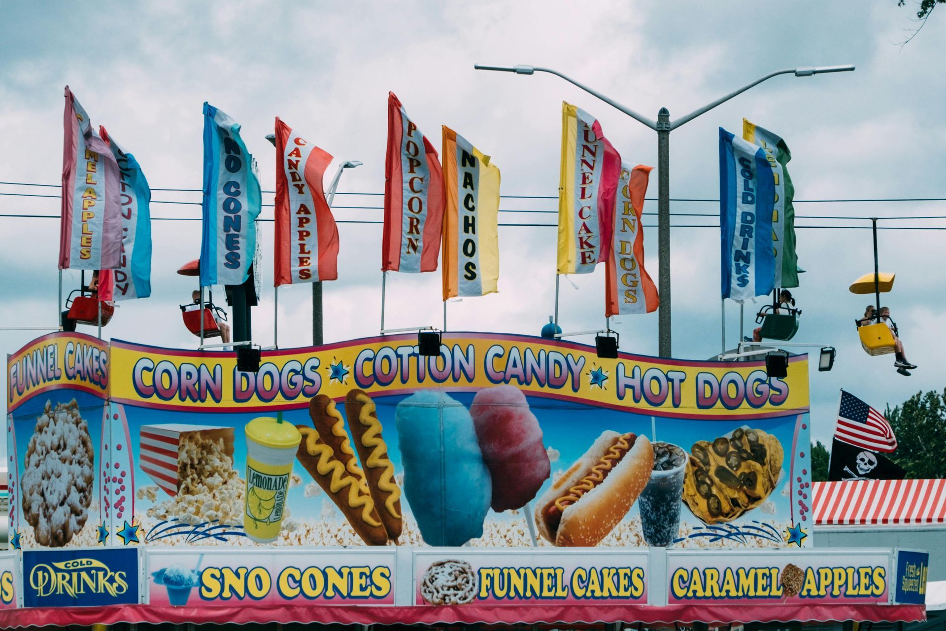 Colorful carnival concession stand with flags and signs for corn dogs, cotton candy, hot dogs, sno cones, and funnel cakes