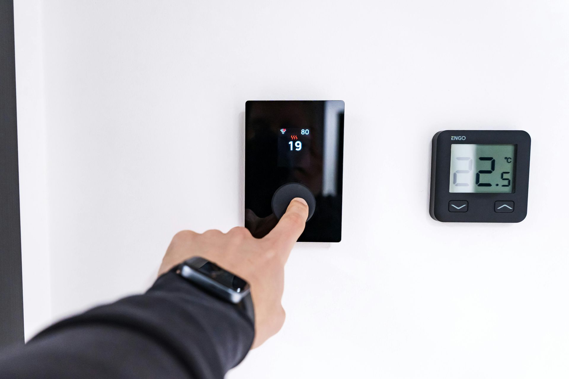 A hand adjusting a black wall-mounted thermostat next to a smaller digital temperature and humidity display.