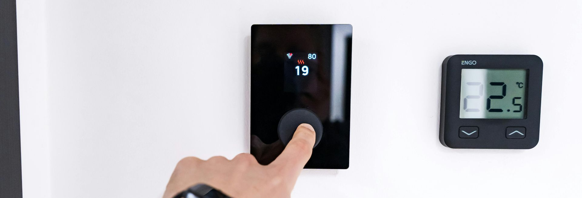 A hand wearing a smartwatch presses a black smart thermostat on a white wall, next to a digital temperature display.