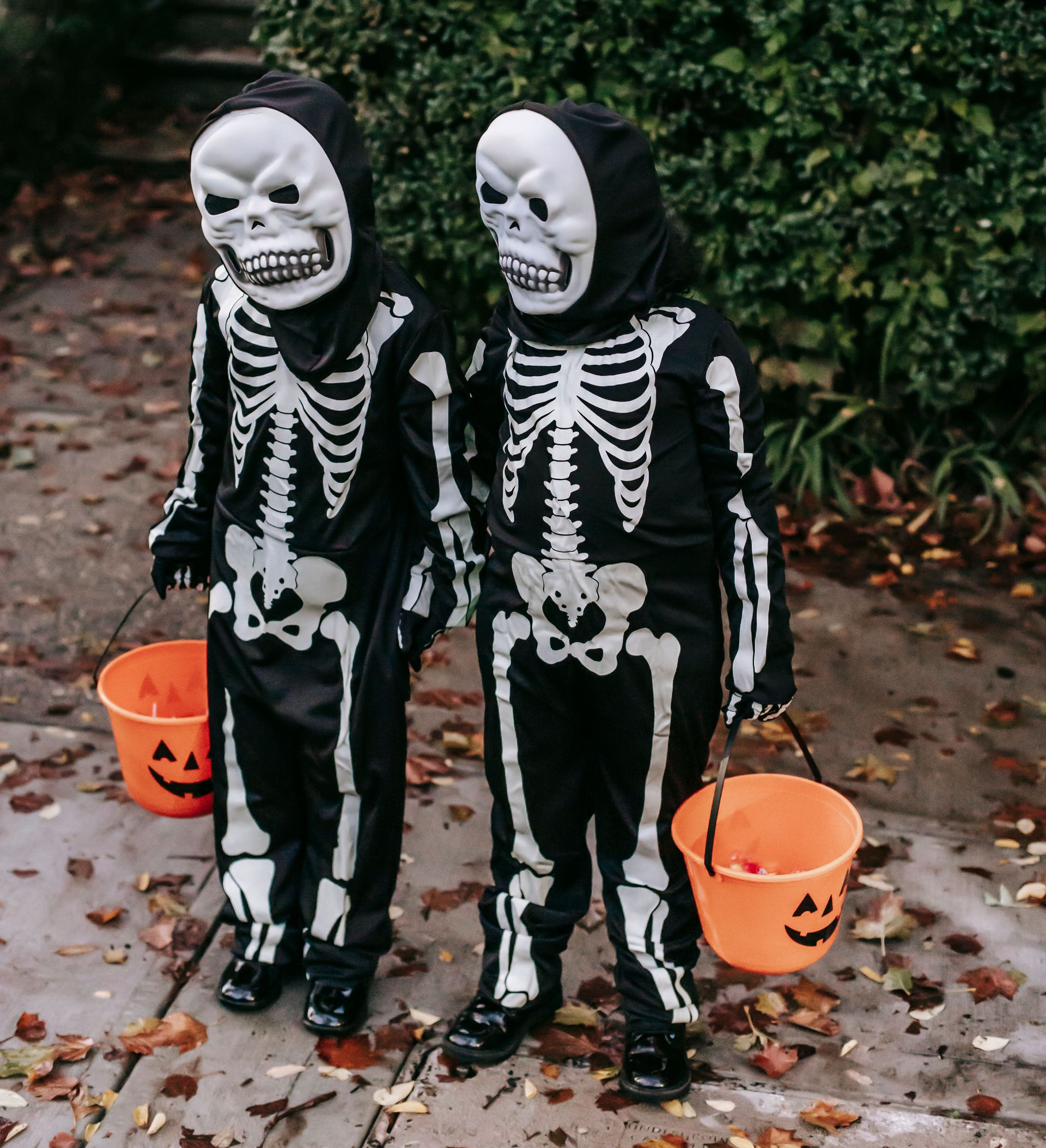 Two children in skeleton costumes holding orange pumpkin buckets on a porch