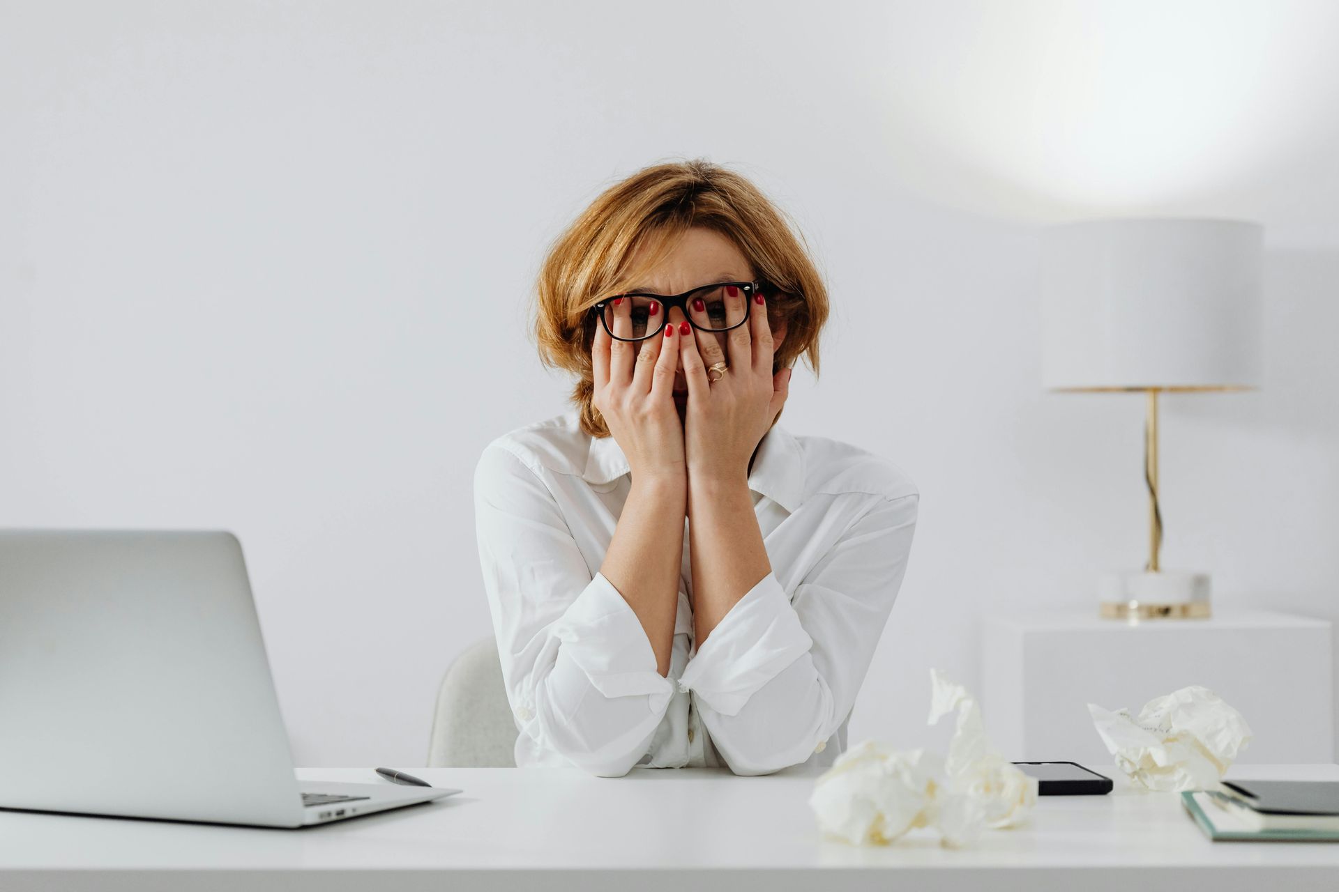 Woman with glasses, covering face with hands at a desk, laptop, crumpled tissues.