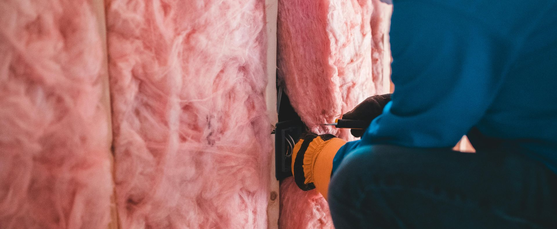 A person in a blue shirt installing pink fiberglass batt insulation between wooden wall studs.