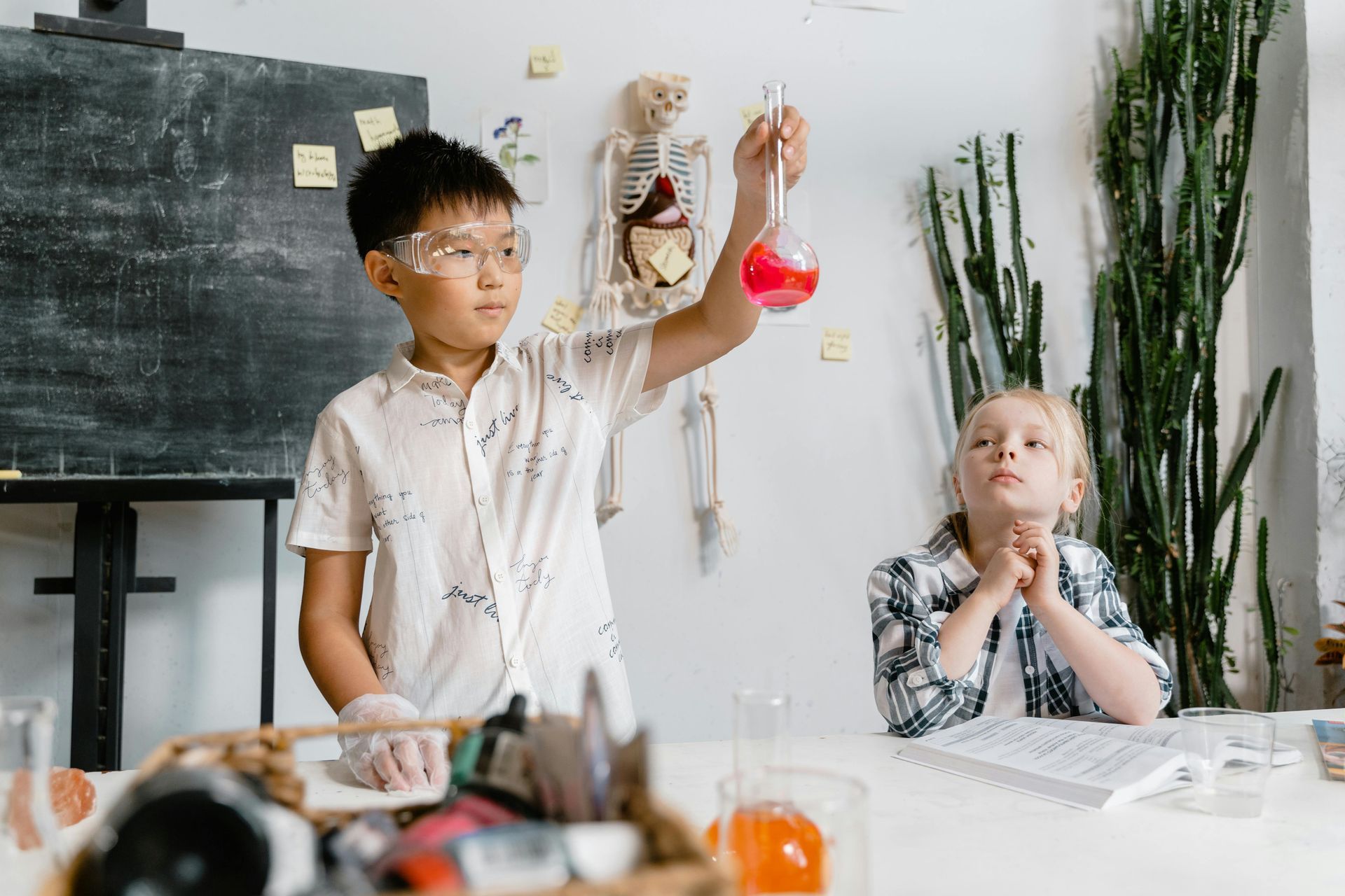 Boy in safety goggles holds a flask of pink liquid as another child watches in a science classroom.