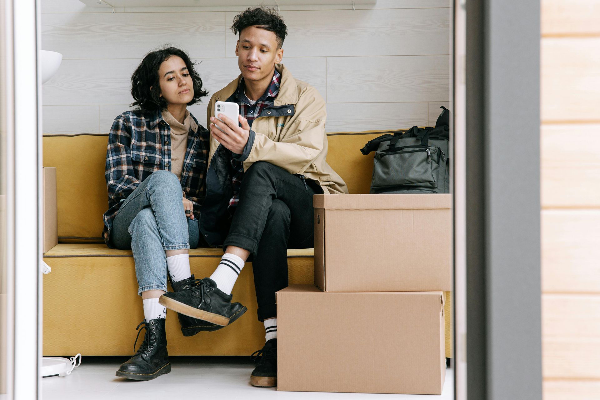 Couple looking at a phone while sitting on a yellow couch next to moving boxes.