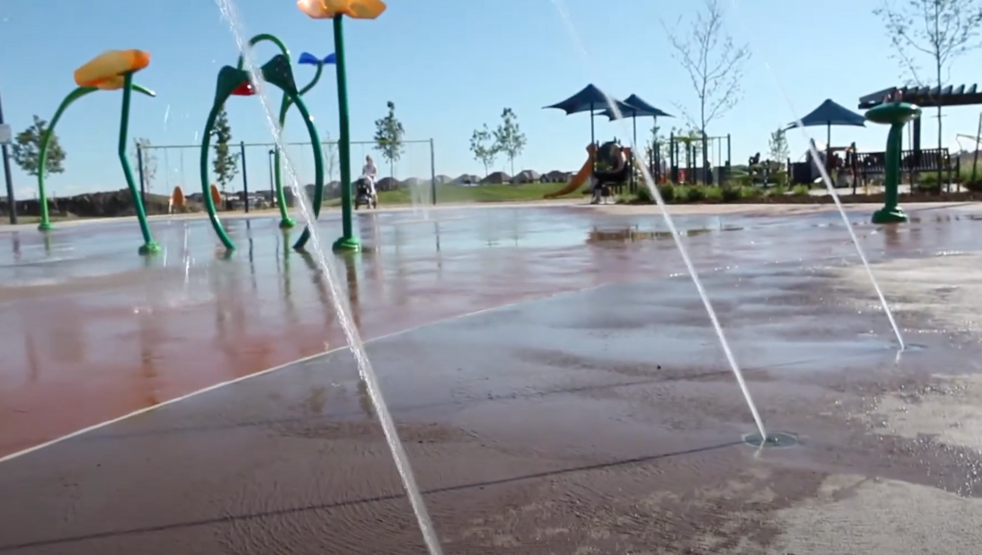 Splash pad with water jets spraying across a wet playground under a sunny sky in Willmott MIlton