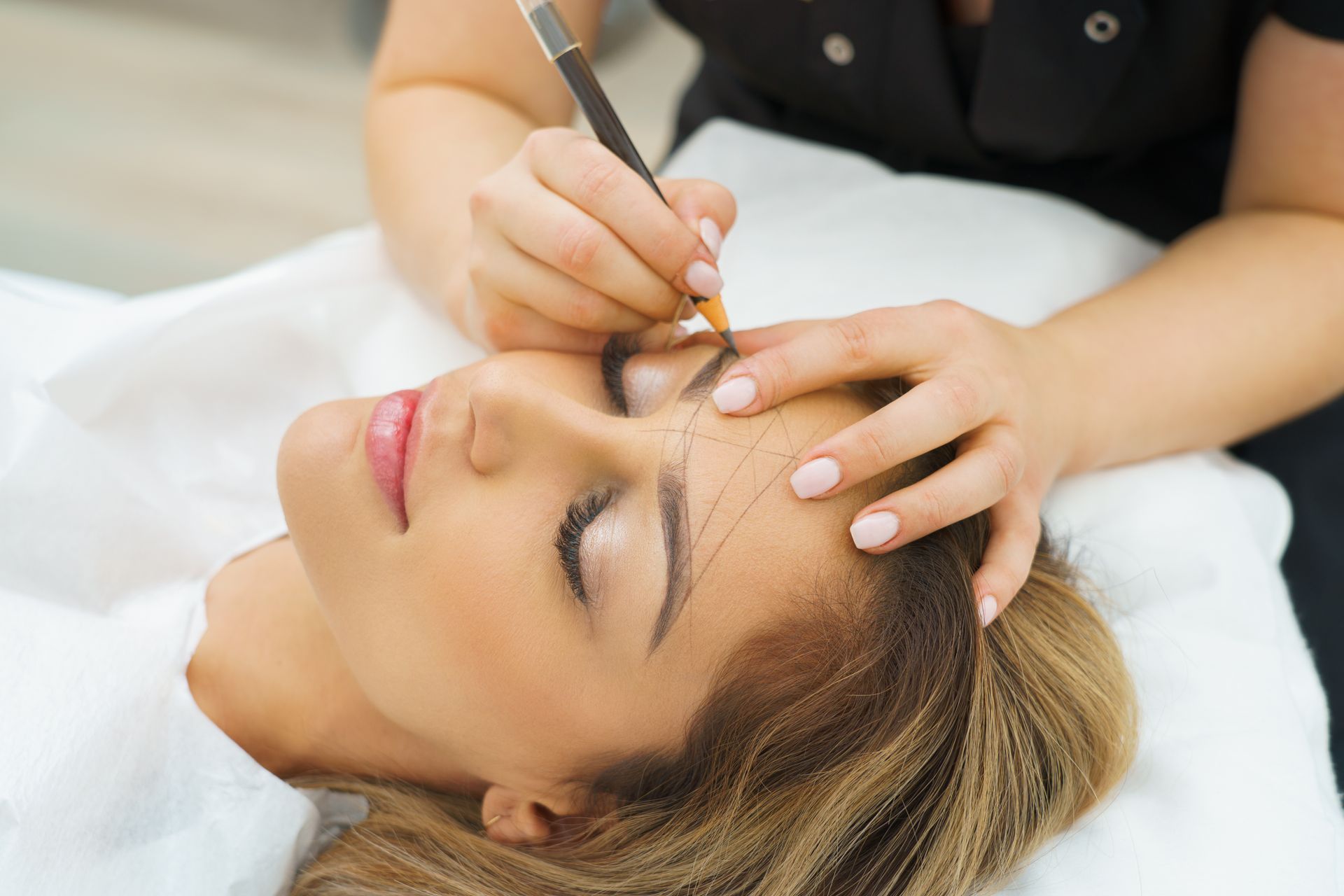 Cosmetician shaping a woman’s eyebrows with a tool while she lies relaxed on a treatment bed