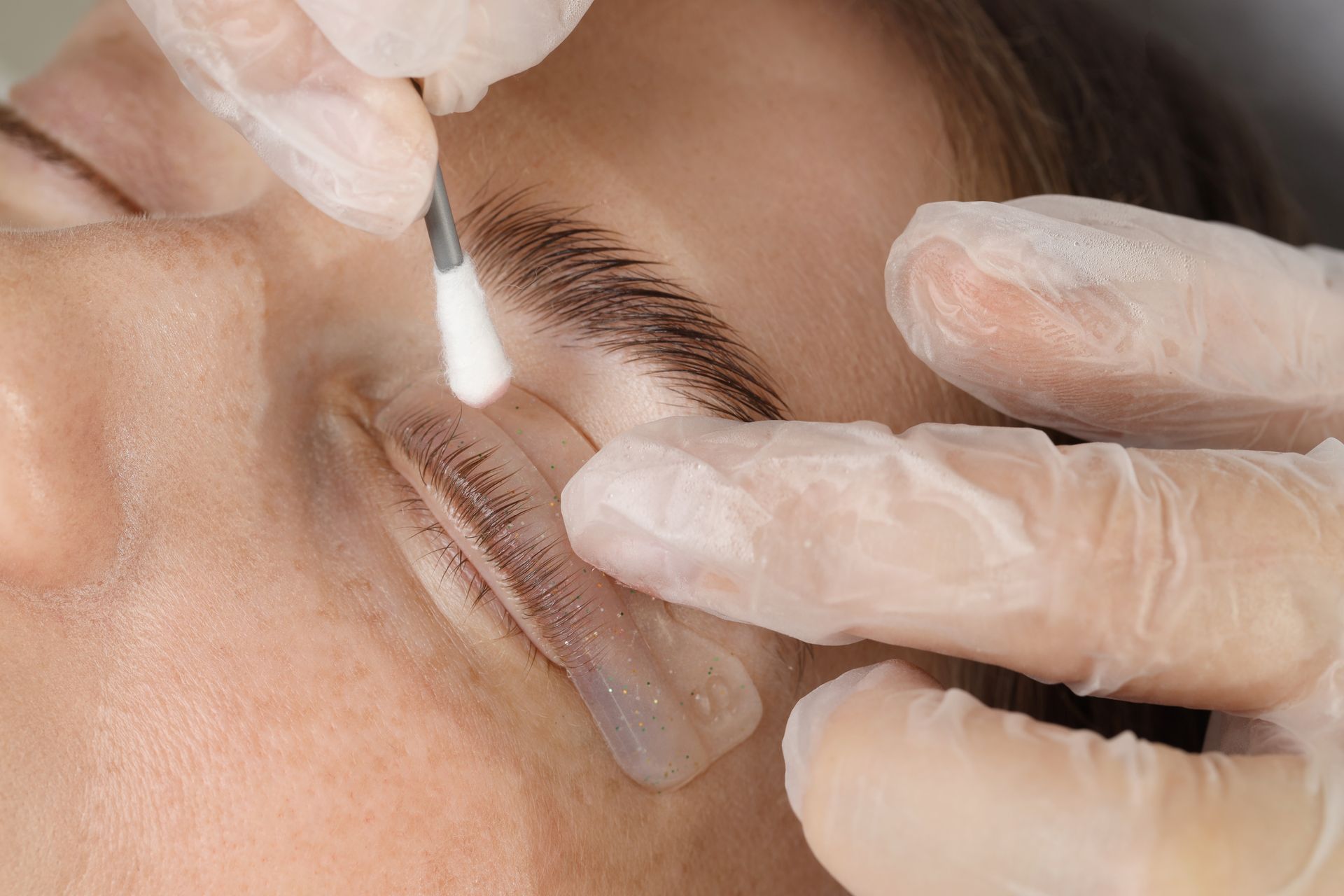 Gloved hands applying eyelash treatment with a brush and pad over a closed eye
