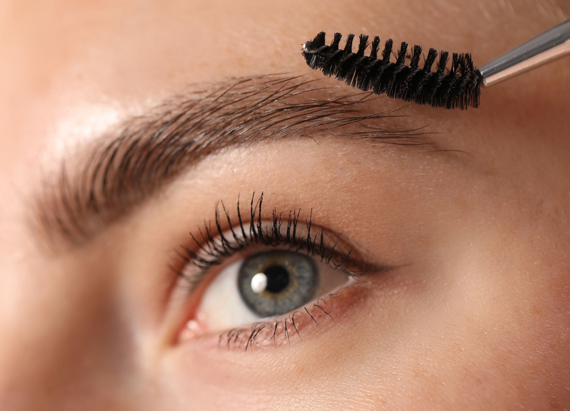 Close-up of an eyebrow being brushed with a spoolie, with one blue eye visible.