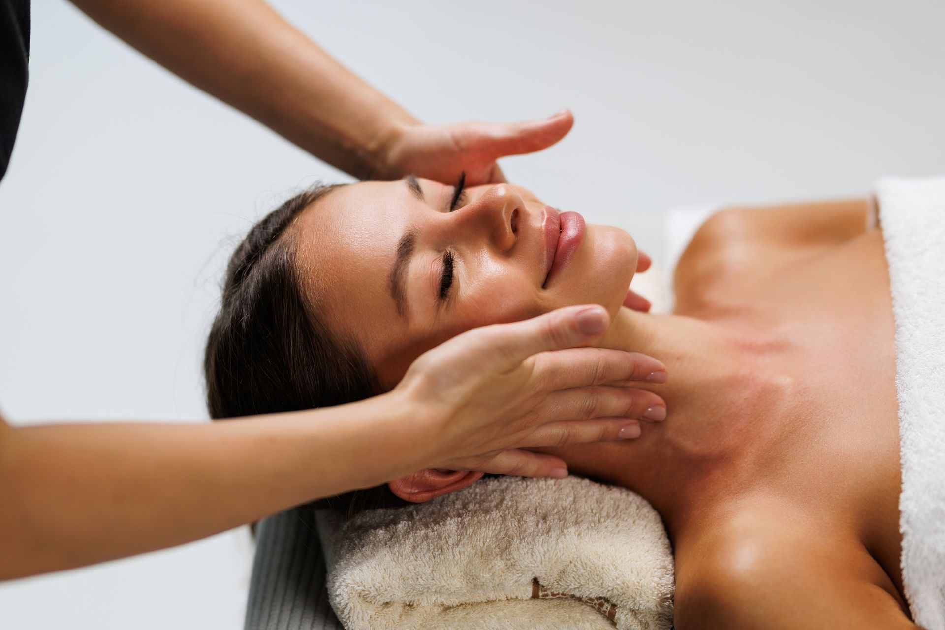 Spa massage with hands gently massaging a relaxed woman’s face and neck on a towel-covered table