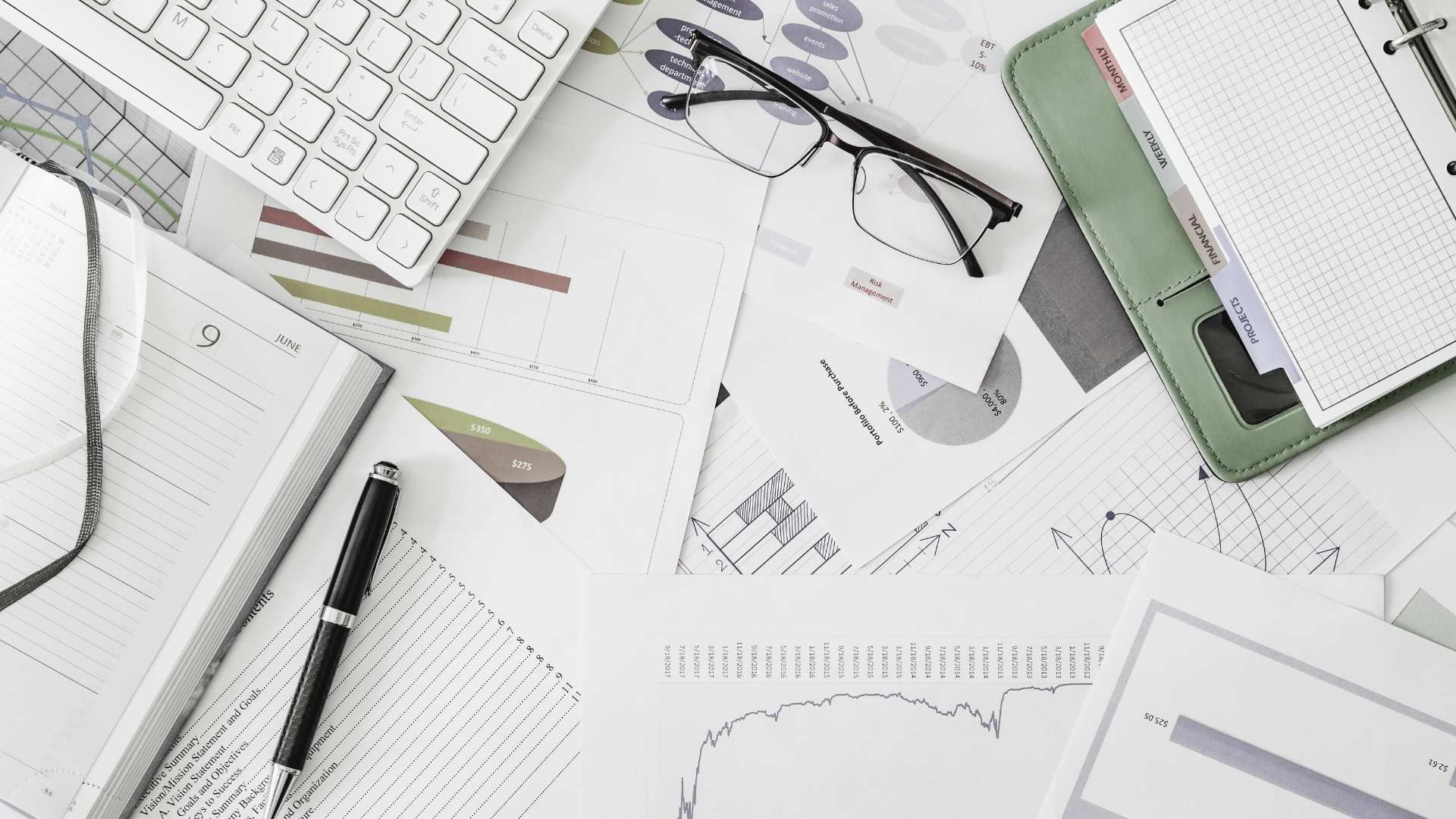 Desk with scattered financial charts, glasses, keyboard, pen, and a green folder on a white workspace