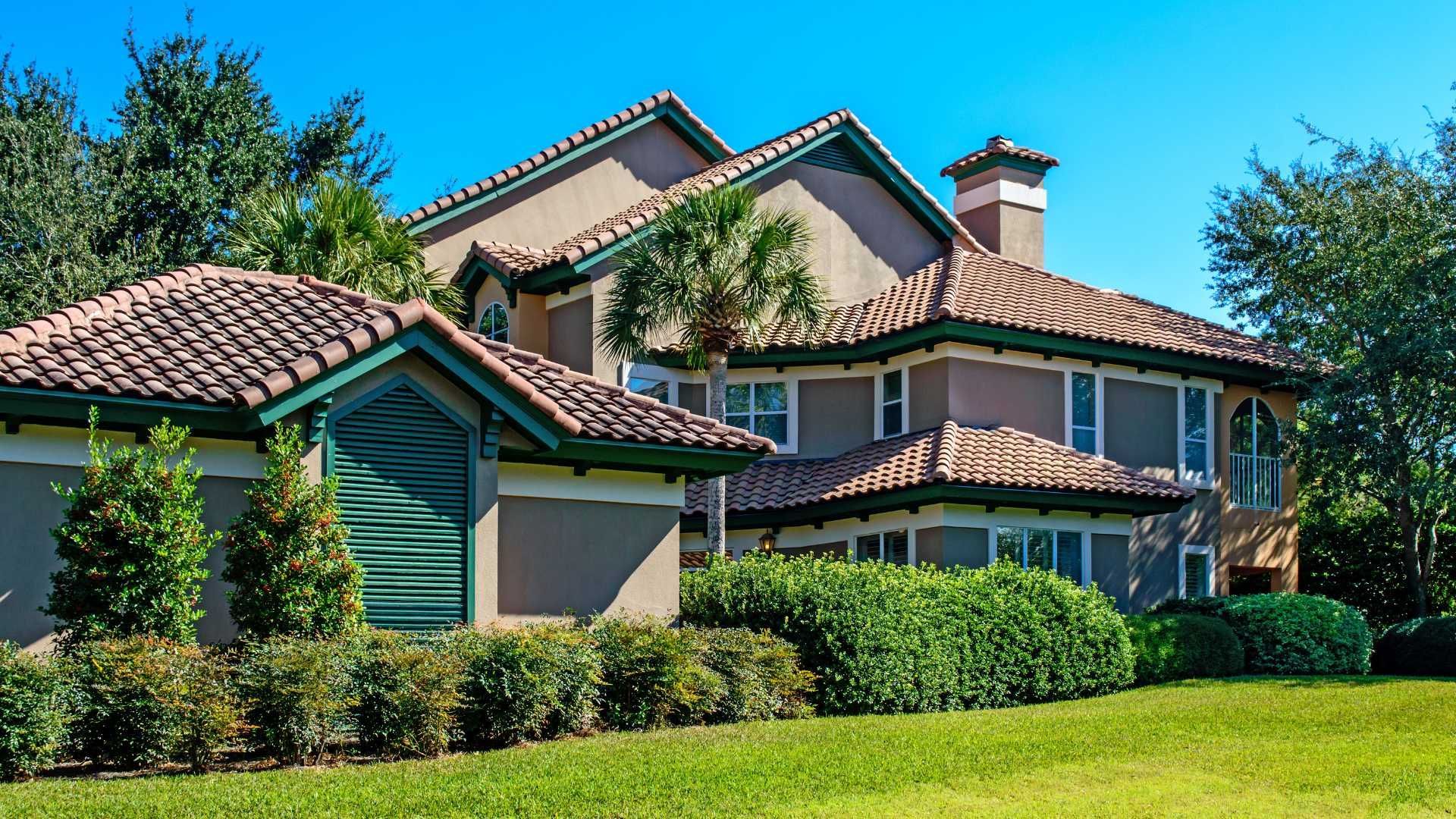 Large beige house with a tiled roof, green shutters, and a manicured lawn under a clear blue sky
