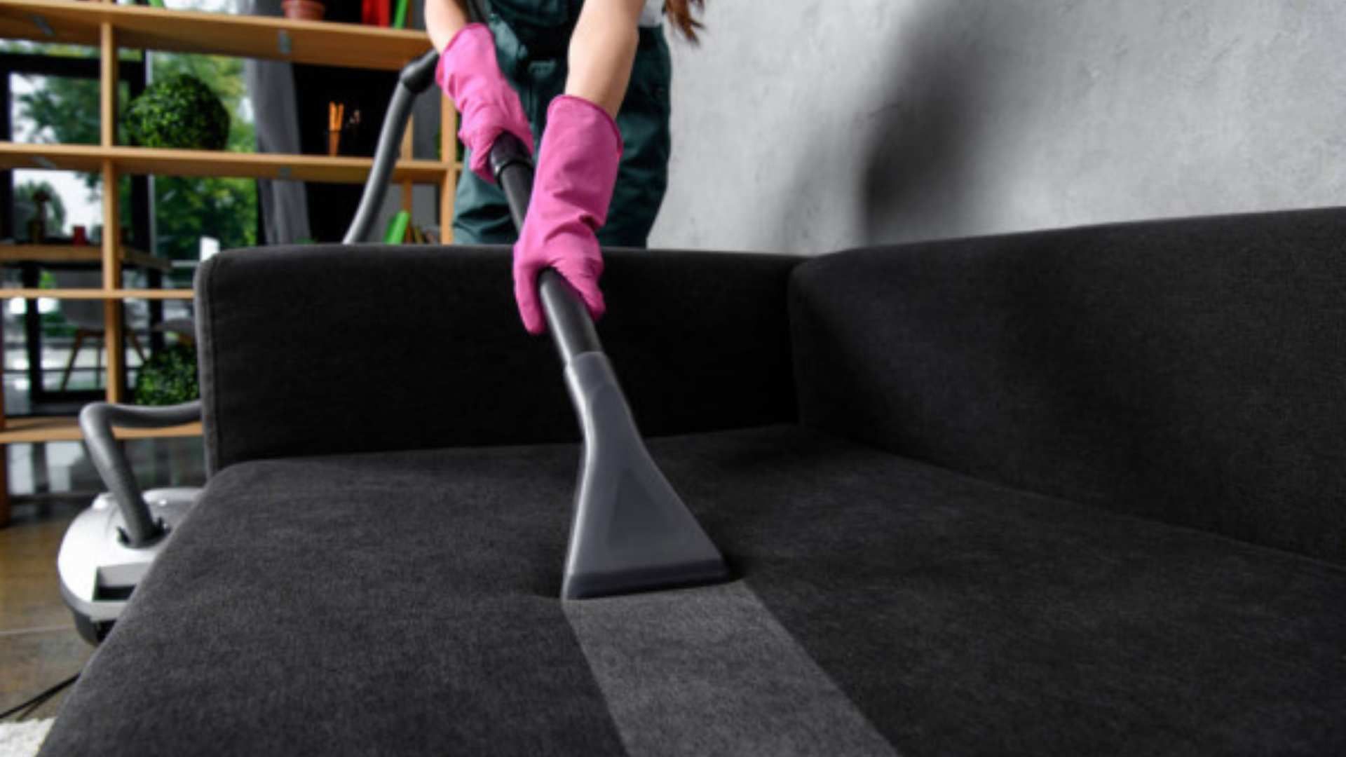 Pink-gloved hands vacuuming a black sofa with a handheld attachment