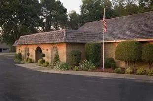 Single-story stone building with arched entry, flagpole, trimmed hedges, and dark roof beside a driveway.