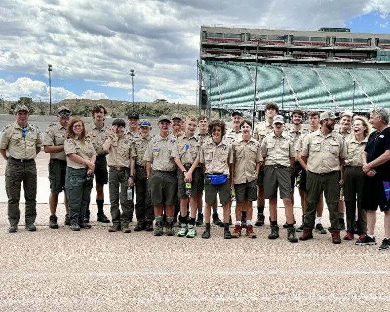 Large group in tan scout uniforms posing outdoors in front of a stadium under a cloudy sky.