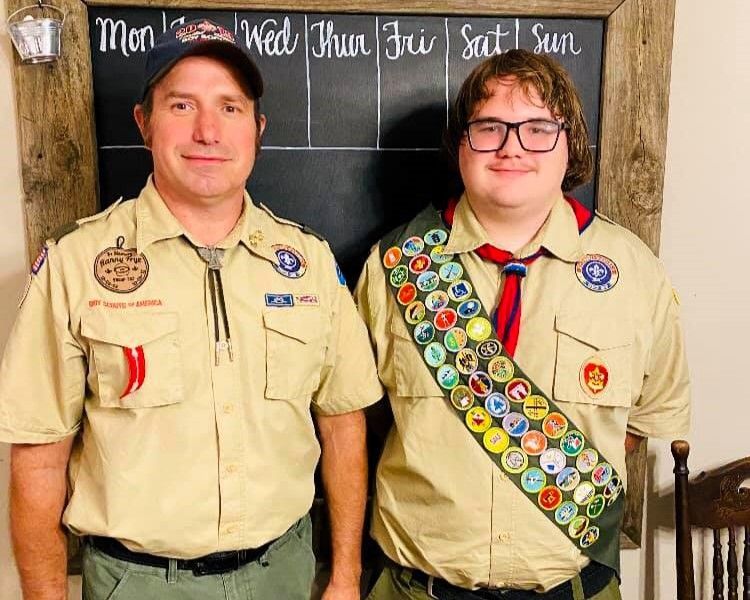 Two Scouts in tan uniforms standing in front of a chalkboard weekly schedule.