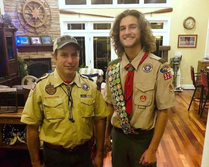 Two scouts in tan uniforms posing indoors, one wearing many merit badges and smiling.