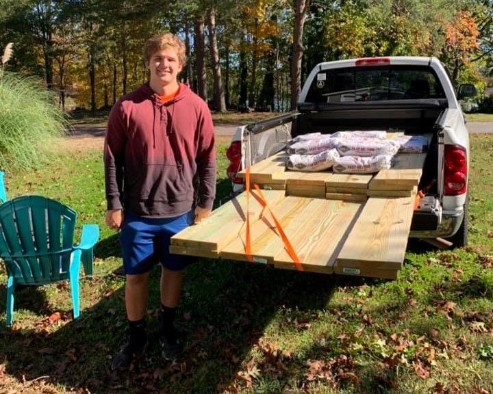 Person stands beside a pickup truck with a wooden table ramp and stacked supplies in a sunny autumn yard