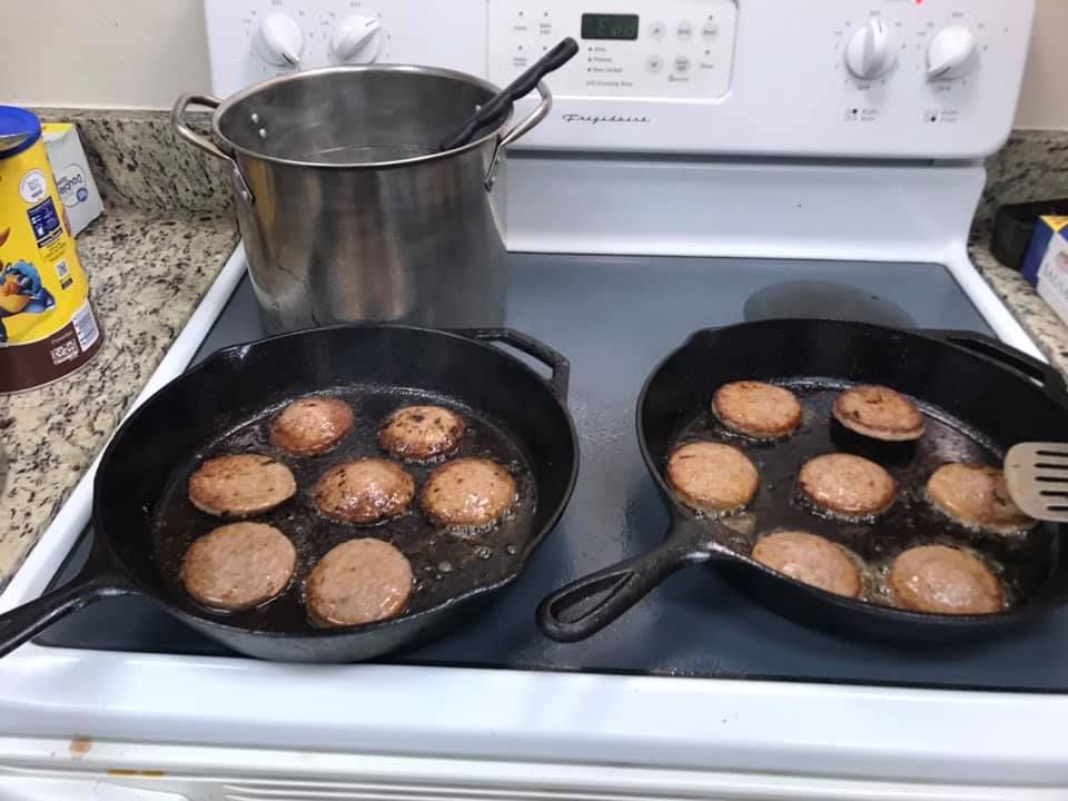 Two pans of frying burgers on a stovetop with a pot simmering on the back burner
