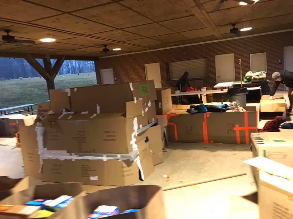 Warehouse room with stacked cardboard boxes, tables, and people working near windows and fluorescent lights