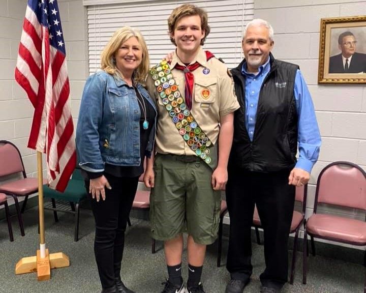 Three people stand indoors beside a U.S. flag, with a Boy Scout in uniform centered between two adults.