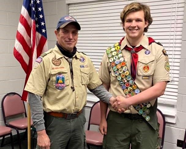 Two scouts in tan uniforms shaking hands, standing indoors beside an American flag.