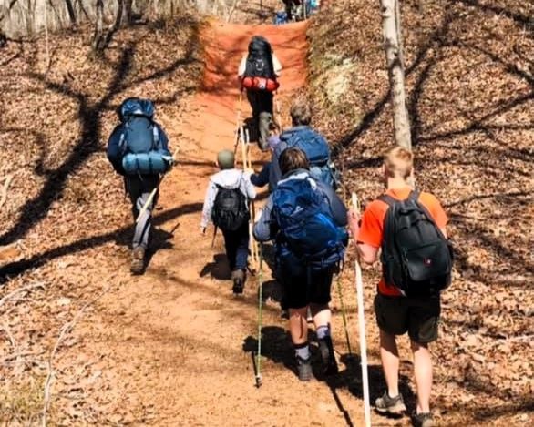Hikers with backpacks and trekking poles climbing a steep dirt trail in a wooded area