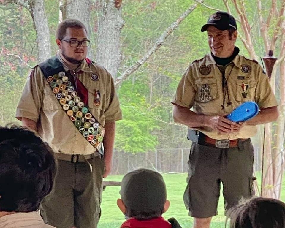 Two scouts in tan uniforms stand speaking to a seated audience under a pavilion.