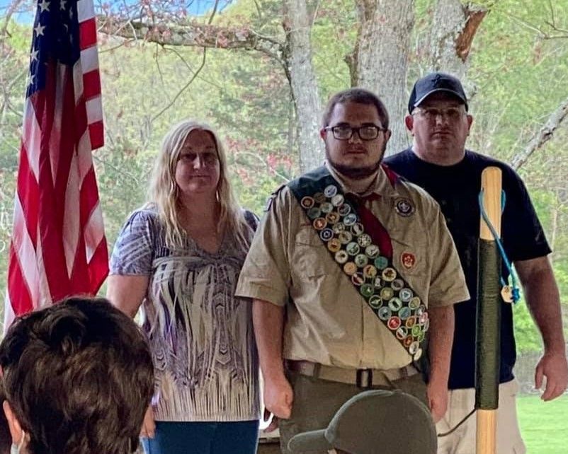Three people standing outdoors; the center person wears a tan uniform with a sash of badges.