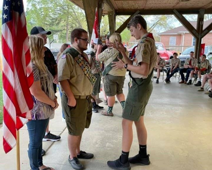 Scout leaders and children chatting under a pavilion, with an American flag at left.