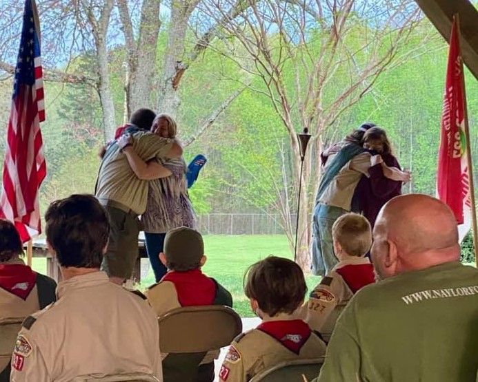 Two soldiers embrace during a ceremony while seated attendees watch under a pavilion, with flags on both sides.
