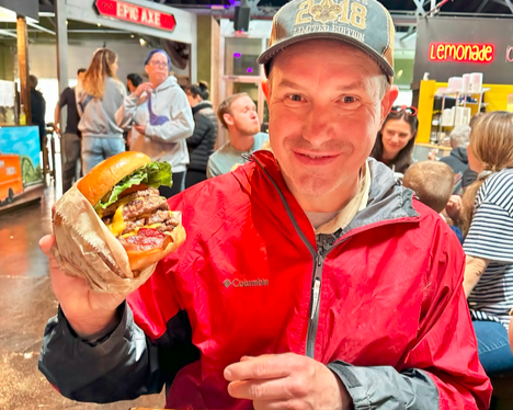 Man in red jacket holding a burger at a busy indoor food market, smiling at the camera