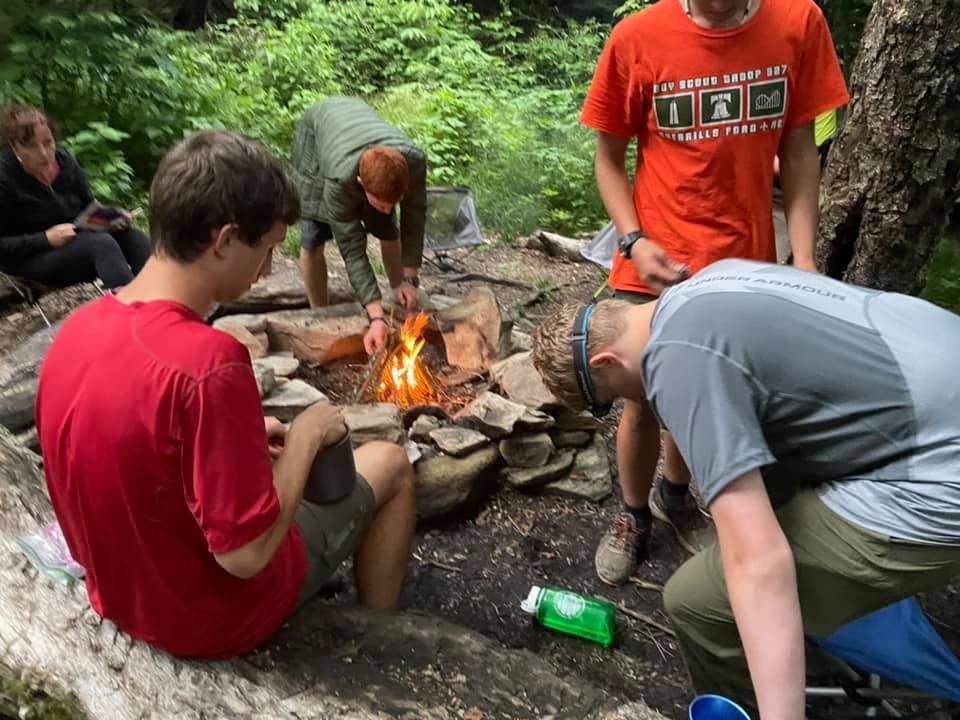 People gathered around a small campfire in a wooded area, cooking or tending the fire.