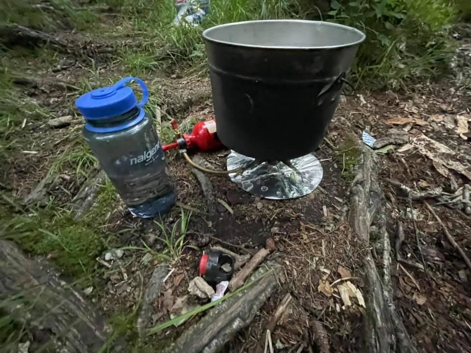 Blue water bottle and black metal pot on a forest floor beside a fire pit.