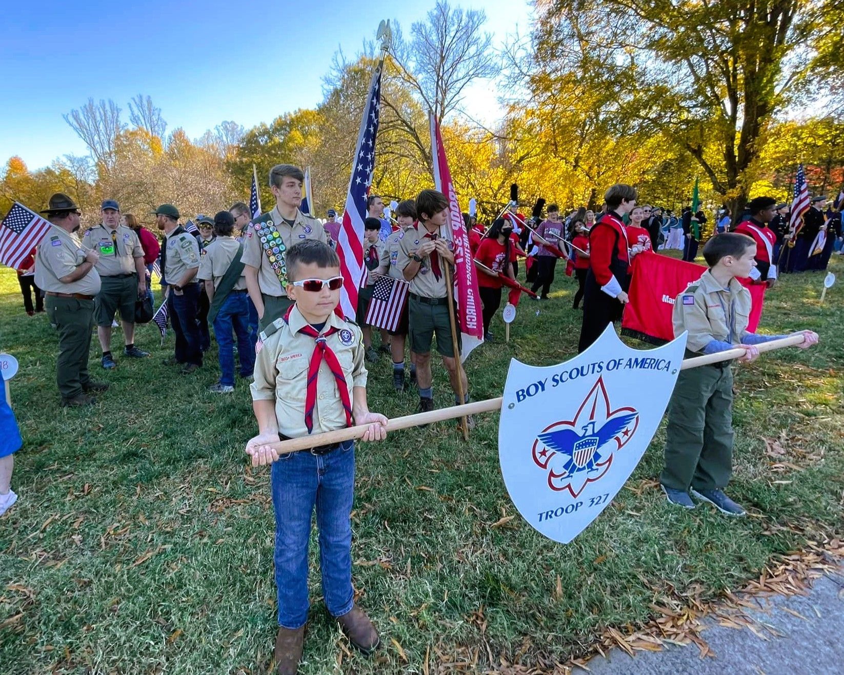 Children in uniforms hold flags at an outdoor parade on a sunny fall day.