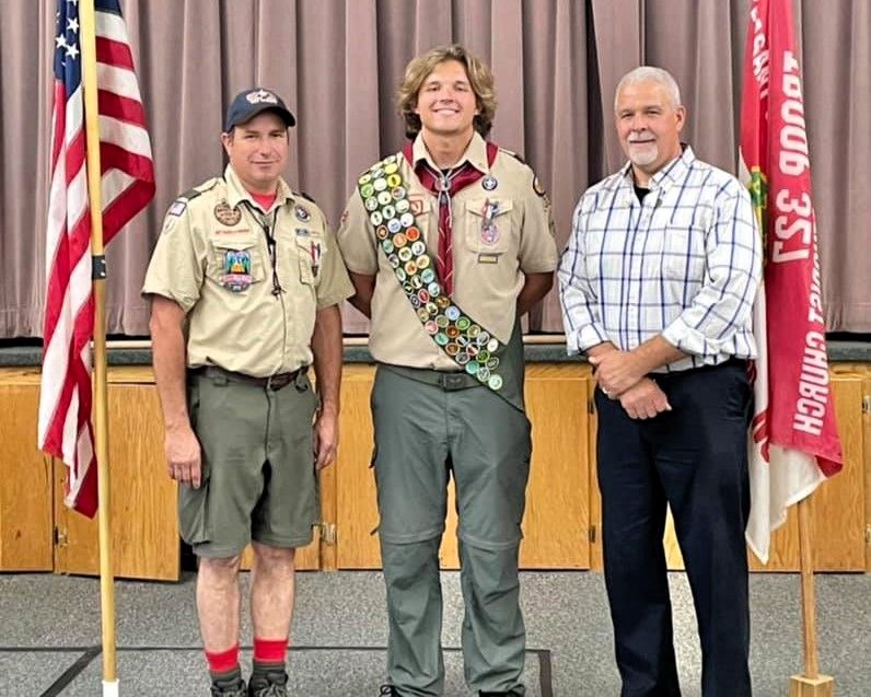 Three men stand on a stage in scout uniforms and casual clothes, with American flags behind them.