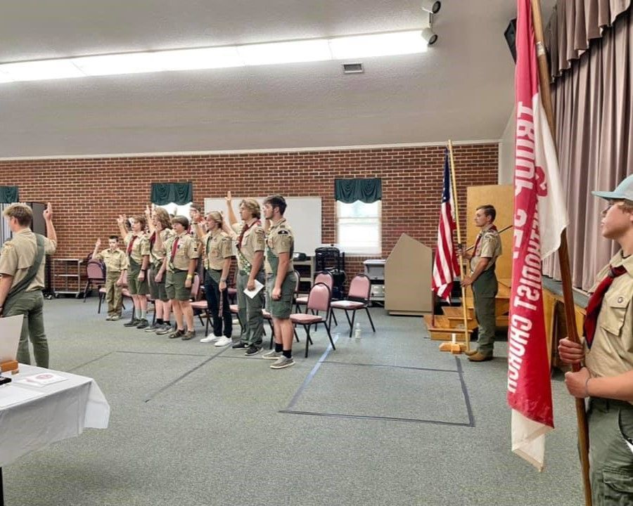 Scouts in uniform lined up on a stage at a ceremony, with flags and brick walls in the hall.