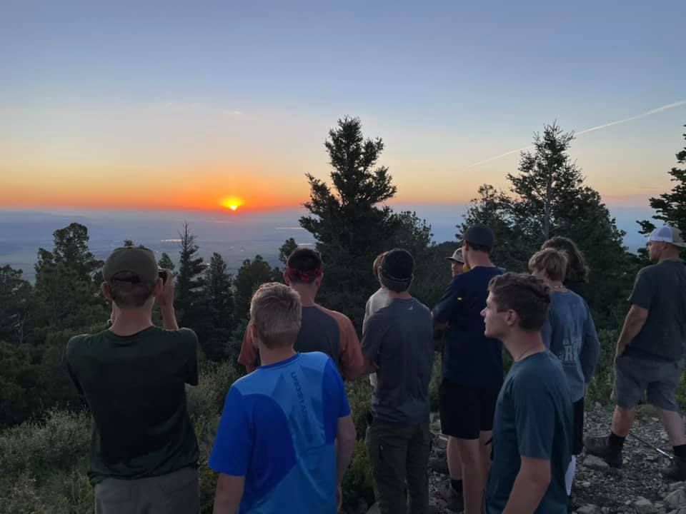 Group of people watching a sunset from a hillside overlook, with trees silhouetted against the orange sky.