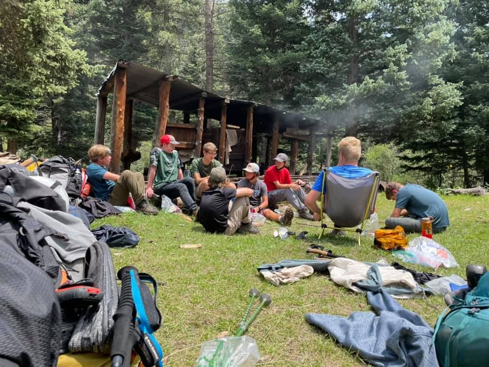 Group of hikers resting on grass beside a forest shelter, with backpacks and gear scattered around.