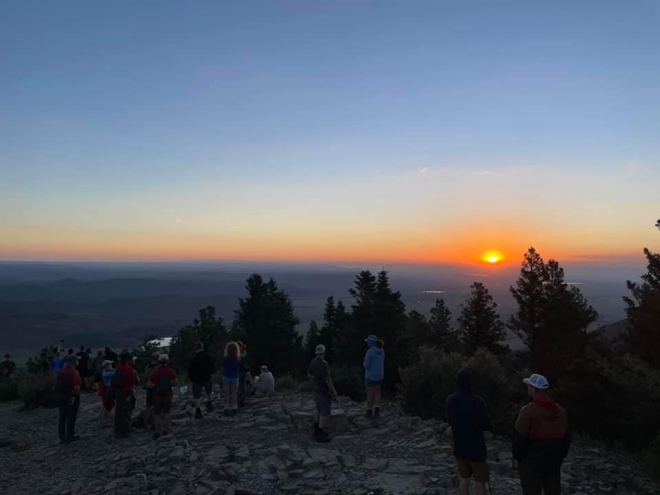 People gathered on a rocky overlook at sunset, with orange glow on the horizon and trees silhouetted in front.