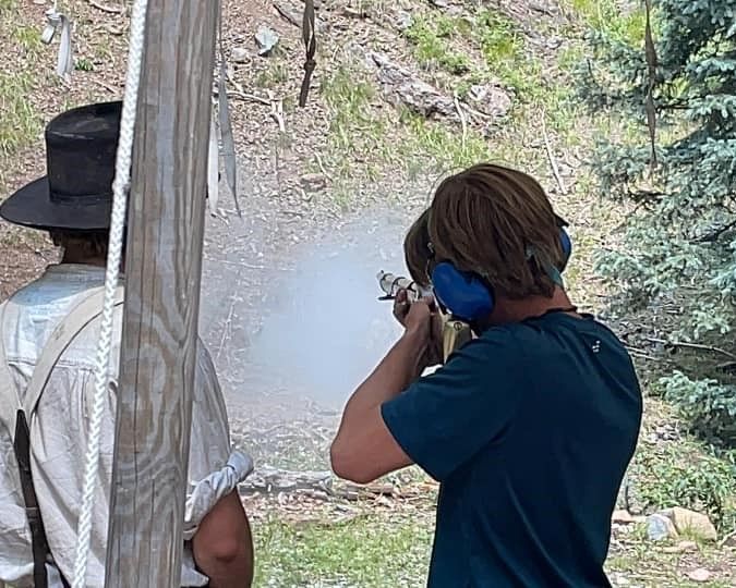 Person aiming a rifle at an outdoor shooting range, with another person nearby under a canopy.