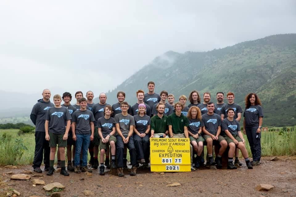 Group of hikers posing outdoors in front of misty mountains, holding a yellow event sign.