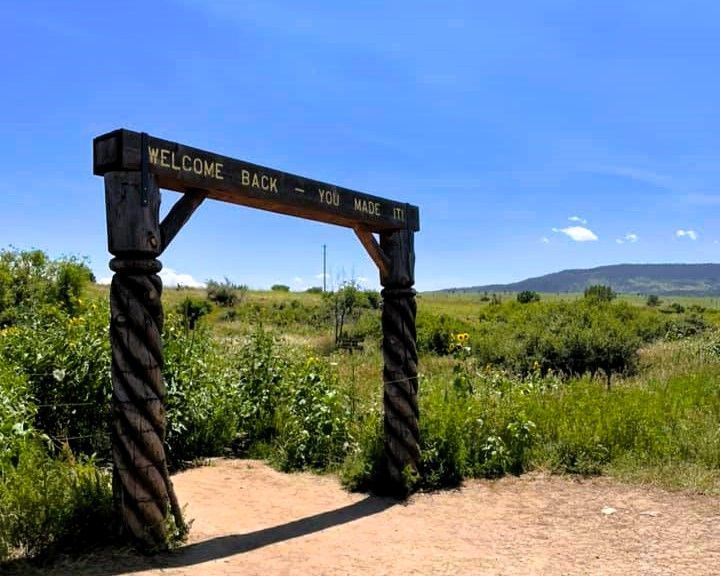 Wooden welcome arch on a dirt path in a sunny grassland with mountains in the distance