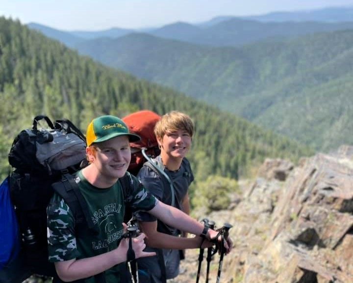 Two hikers with backpacks smiling on a mountain ridge with forested valleys behind them