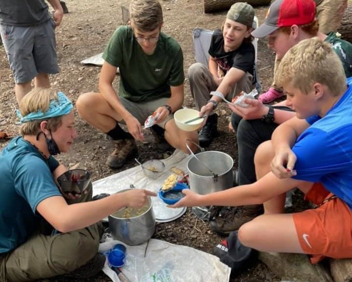 Kids sitting outdoors around metal buckets, scooping and pouring liquid in a group activity