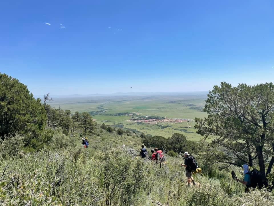 Hikers descending a grassy hillside with trees, overlooking a wide green valley under a blue sky