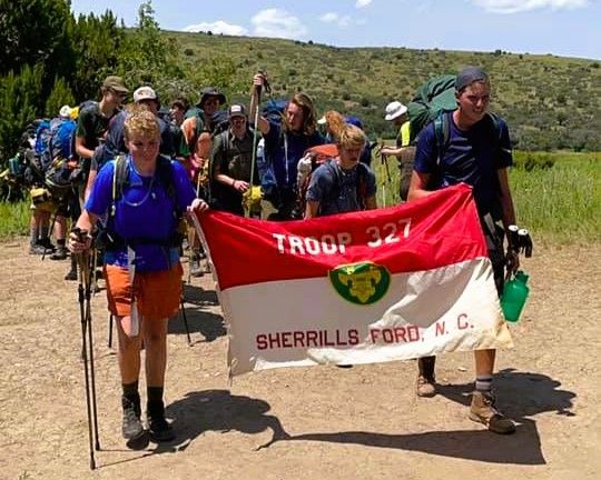 Scouts carrying a red Troop 327 banner on a sunny hiking trail in a grassy hillside.