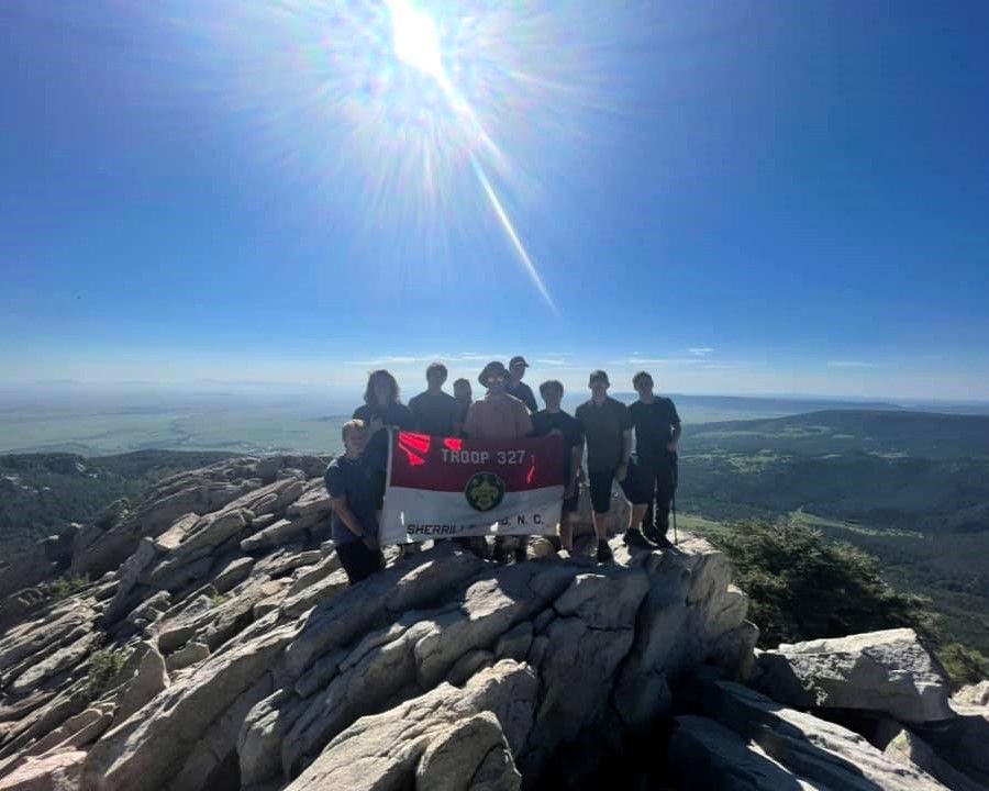 Group of hikers on a rocky mountain summit holding a banner under a bright blue sky.