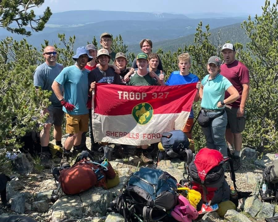 Hikers posing with a Troop 327 Sheriffs Ford flag on a mountain overlook with backpacks in front