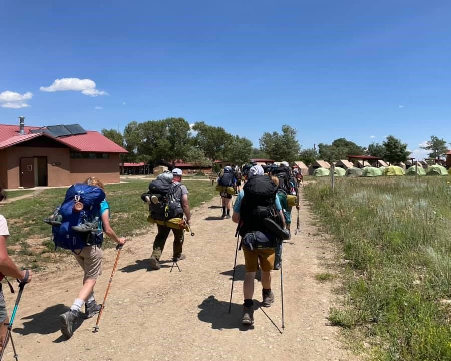Hikers with backpacks walk along a dirt path past a red-roofed building and grassy field under a blue sky.