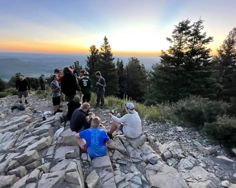 Group of hikers resting on rocky summit at sunset, with trees and distant mountain view.